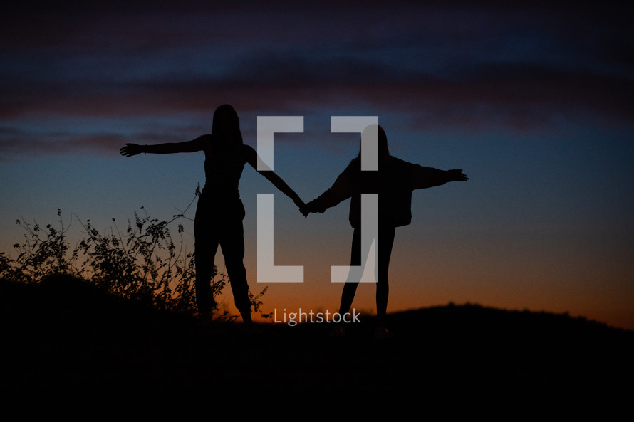 silhouettes of young women on a mountaintop at sunset with hands raised 