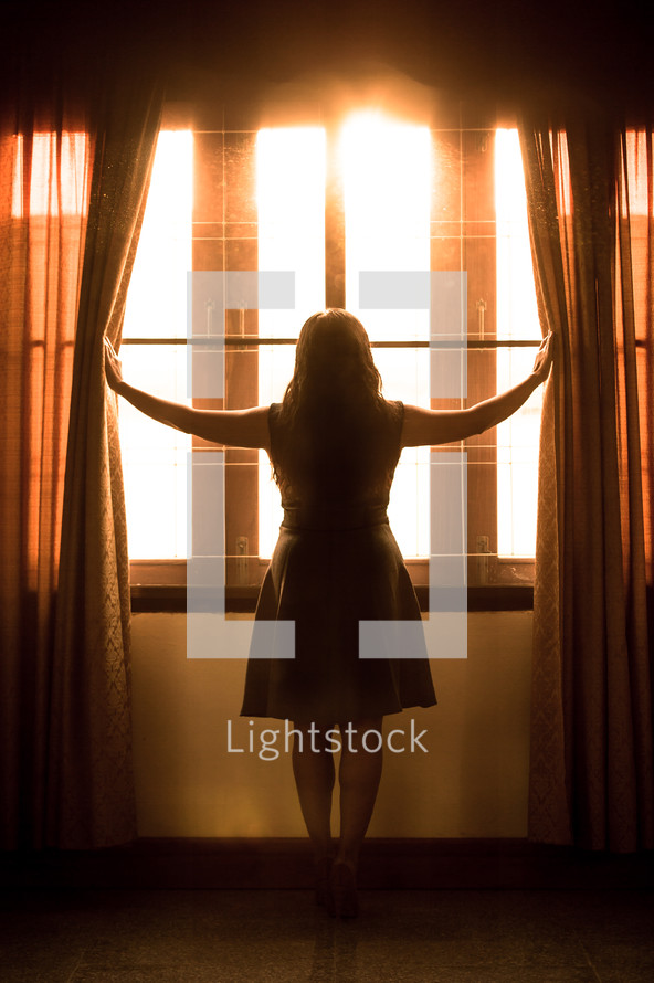 a woman pushing open curtains to look out a window 