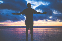 a man standing on a beach on wet sand with open arms at sunset 