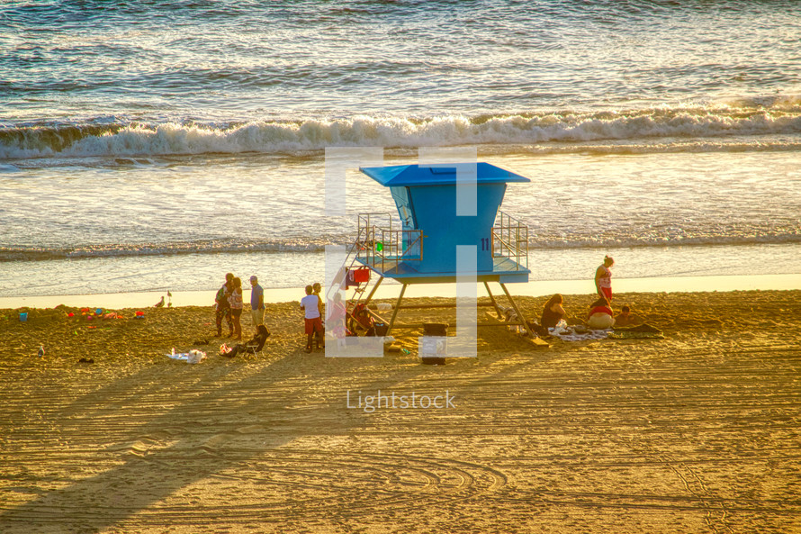 A group of people on a beach sitting around a lifeguard stand at sunset 