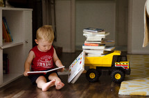 a toddler boy sitting and reading books next to a toy dump truck 