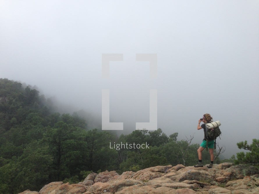 Hiker standing on a rocky mountaintop looking through binoculars into a valley of tree tops.