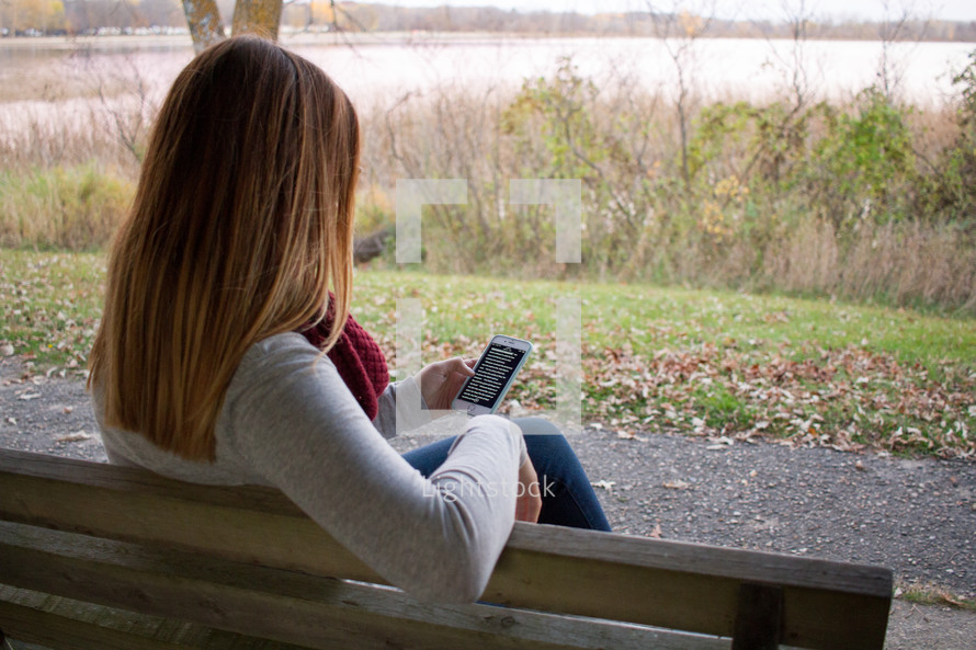 a woman reading a Bible app on her cellphone sitting on a park bench 