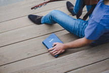 man with a guitar sitting on wooden steps next to a Bible 