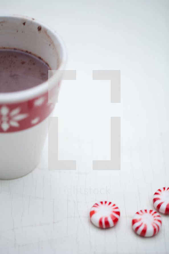 A cup of hot chocolate in a Christmas cup and peppermint candies on a white table.