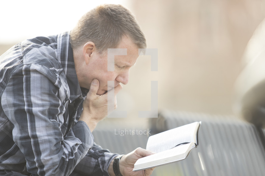 a man in a plaid shirt sitting on a bench outdoors reading a Bible 