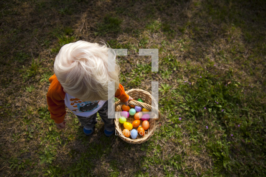 Child carrying a basket of Easter eggs while standing in the grass.