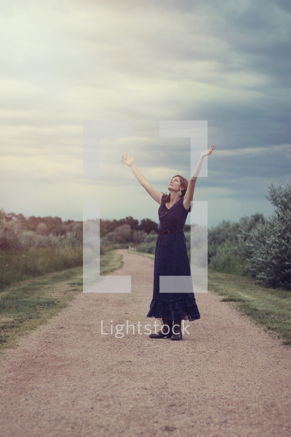 woman standing on a dirt road with raised arms in worship 