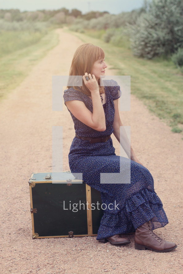 woman sitting on a dirt road next to luggage 