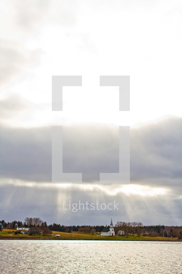Rays of light shining through storm clouds over a hillside church and lake.