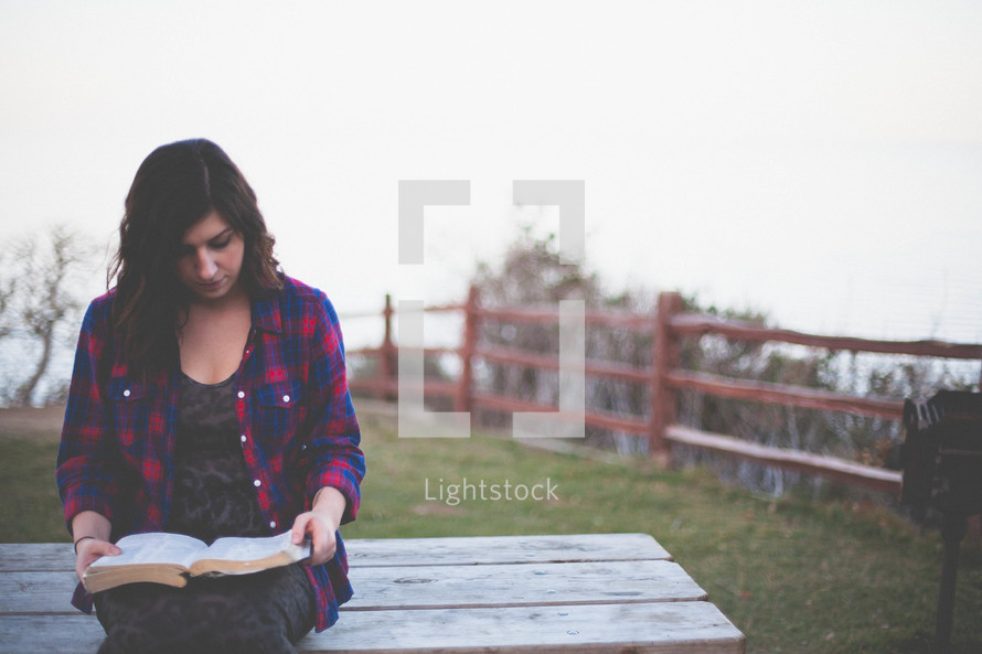 Woman reading the Bible while sitting on an outdoor picnic table.