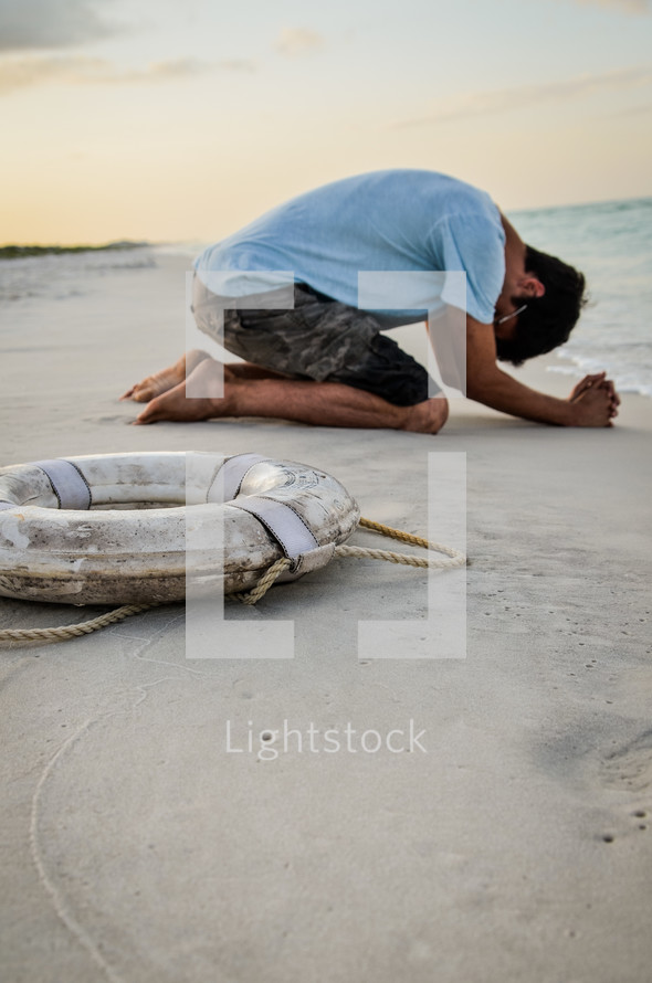 man kneeling in prayer on a beach and a life ring