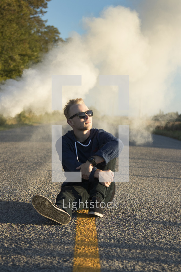 a man sitting in the middle of a road with smoke behind him 