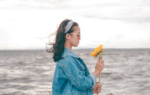 a young woman holding a sunflower standing on a beach 