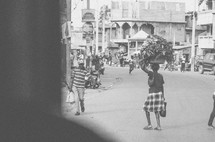 Pedestrians on a village street. Woman carrying basket of leafy produce on her head.