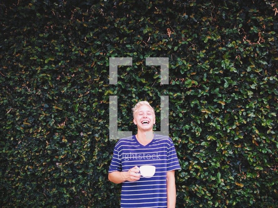 A man standing in front of an ivy covered wall drinking coffee. 