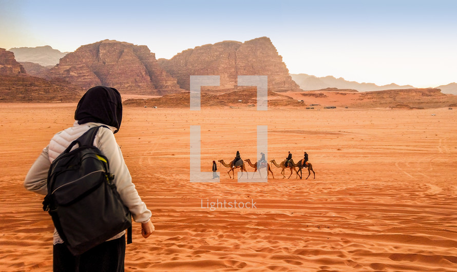 Jordan, Wadi Rum. Caravan of camels with drovers in the desert, rock mountains at sunset. Tourist from behind looks at the panorama.