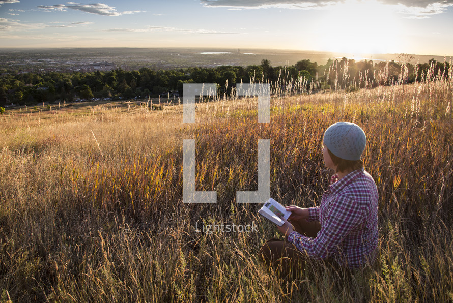 a man reading a Bible in tall grass at sunrise 