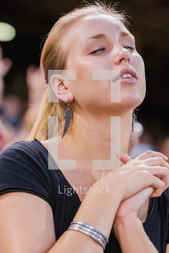 Close up of woman with eyes closed and face towards the sky with hands in praying position with other peopel behind her.