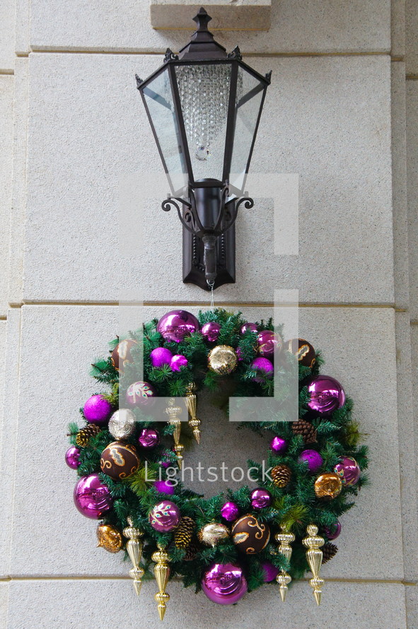 Purple and gold ornaments on pine wreath hanging under coach lamp on stone wall.