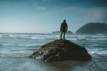 a man standing on a rock on a beach looking out at the ocean 