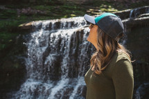 a woman in a ball cap standing outdoors by a waterfall 
