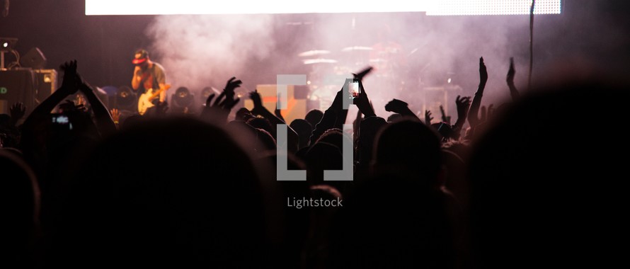 Crowd of people facing hazy stage watching concert with arms raised in the air.