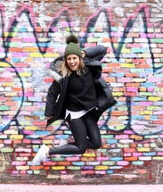 young woman jumping up in front of a graffiti covered wall 