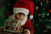 Cute little boy in Santa Claus costume eating gingerbread house on Christmas tree background. Home celebration, decoration with lights. Happy childhood, kids, lovely son. High quality photo