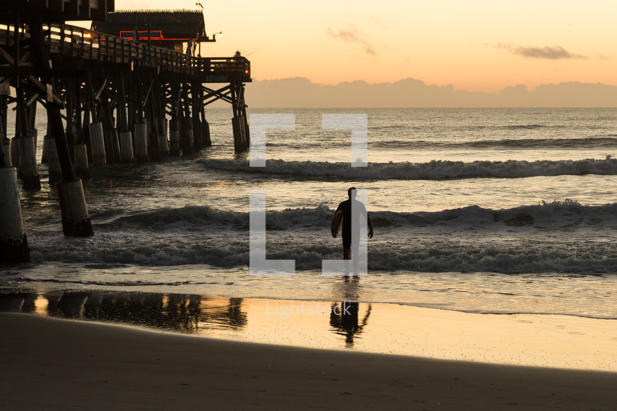Silhouette of a man carrying a surfboard walking out in to the ocean waves at daybreak near a pier.