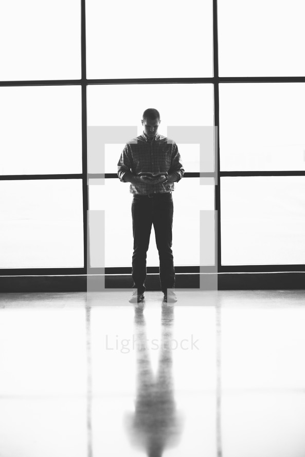 man reading a Bible standing in front of windows 