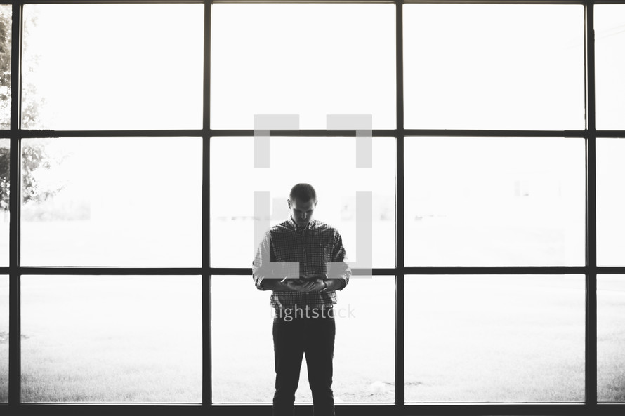 man reading a Bible standing in front of windows 