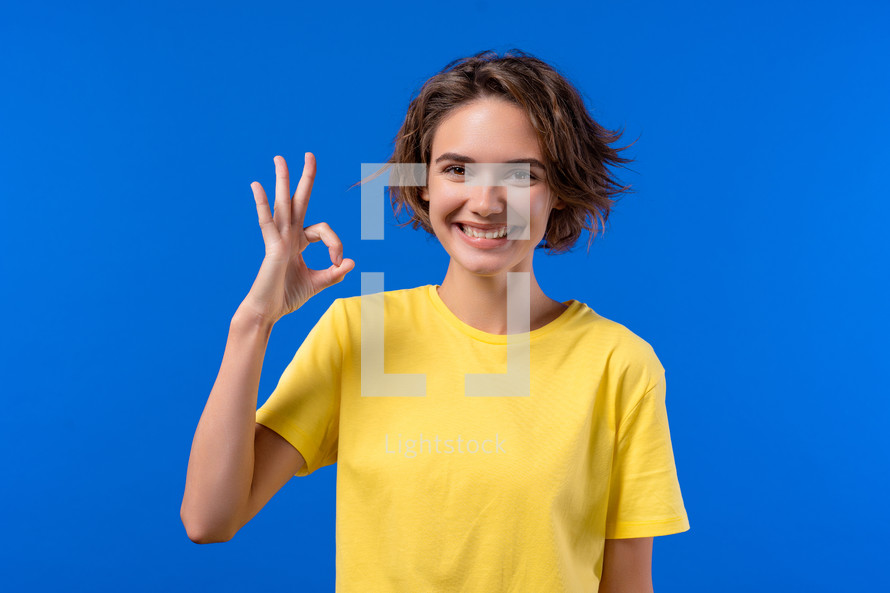 Pretty woman makes hand sign okay, ok gesture. Happy girl, correct perfect choice, great deal, blue background. Positive female model smiles to camera, approval, trust concept. quality
