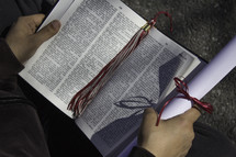 student reading a Bible holding a diploma and graduation tassel