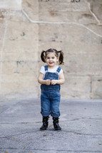 A little girl in overalls in front of a stone wall.