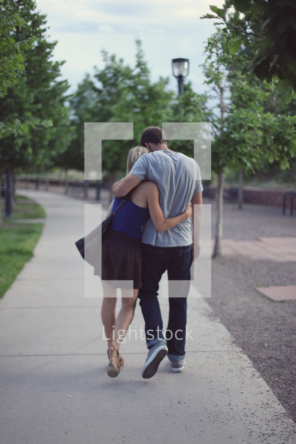 Man and woman embracing as they walk along sidewalk through park.