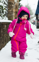 Adorable toddler baby girl in a magenta snow suit playing on the snow.