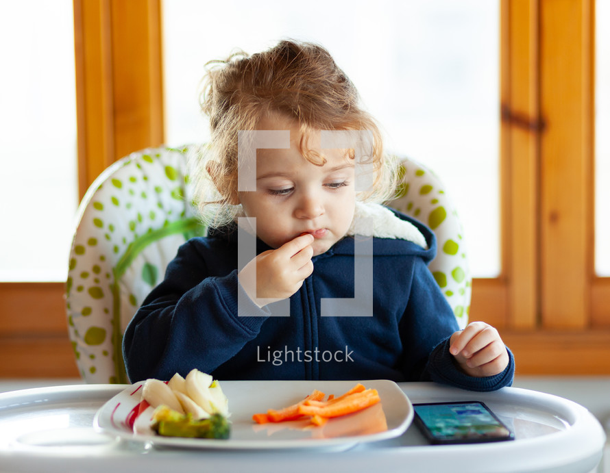 Toddler eats in the high chair while watching movies on the mobile phone. 