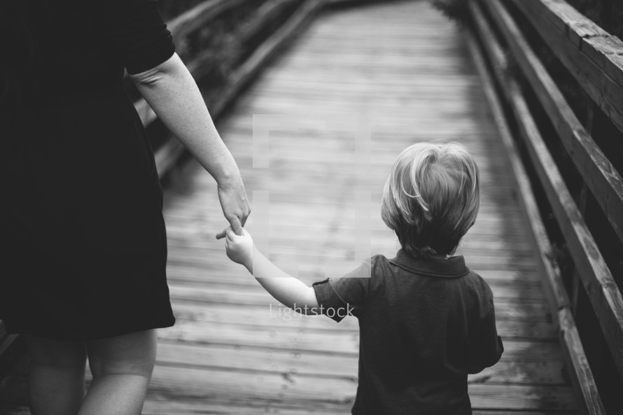 Mother holding child's hand as they walk across a wooden bridge.