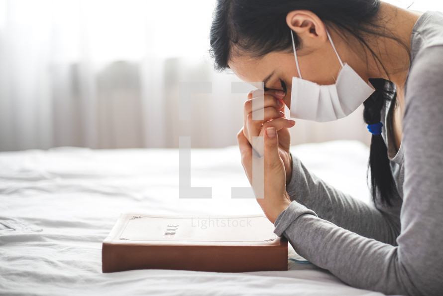 Woman with mask praying next to bed with her bible near.