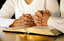 A married couple wearing white shirts clasp their hands in prayer together over an open Holy Bible.  Main focus point is on the woman's hands.