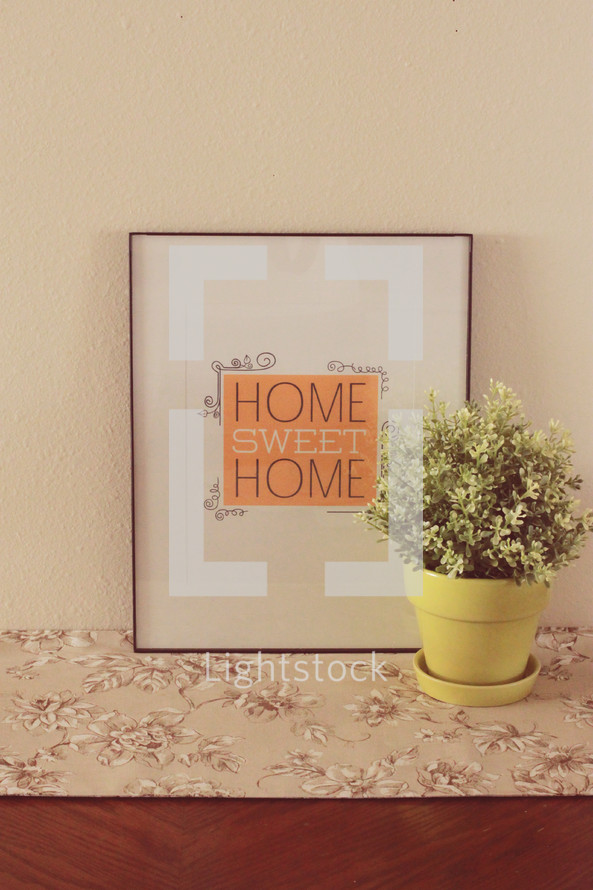 A framed picture reading "Home Sweet Home" next to a pot of flowers.