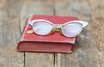 Eye glasses on an old red book, which is on a rugged wooden table.