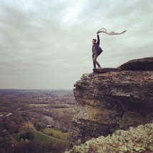man holding a scarf blowing in the wind standing at the top of a mountain 