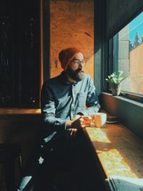 man with a wool cap and beard drinking coffee looking out a window 