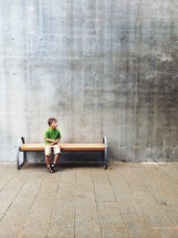 Boy sitting on a park bench against a cement wall.