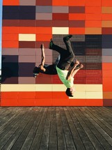 Two men doing back flips in front of a colorful wall.