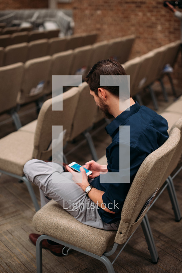 a man sitting in a chair at church looking at his cellphone 