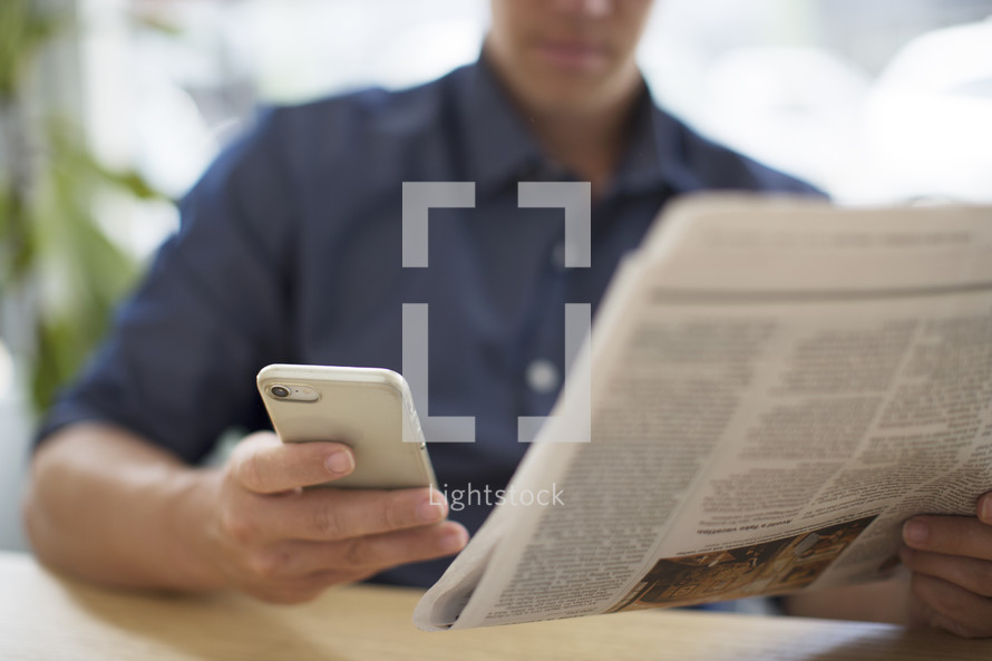 a man reading a newspaper and looking at his cellphone 