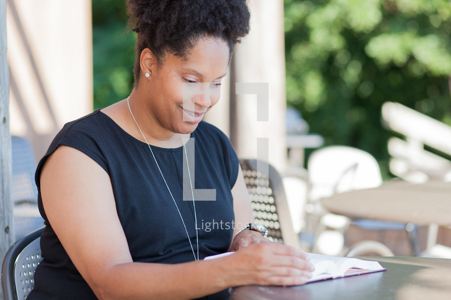 an African American woman sitting at a table reading a Bible outdoors 